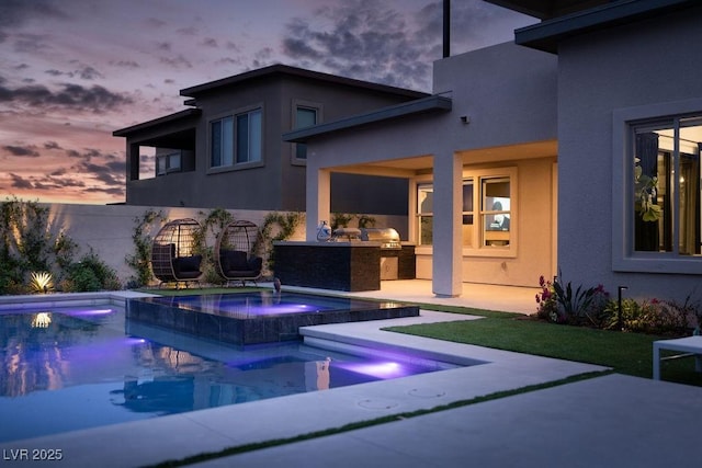 back of house at dusk featuring exterior kitchen, an outdoor pool, an in ground hot tub, and stucco siding