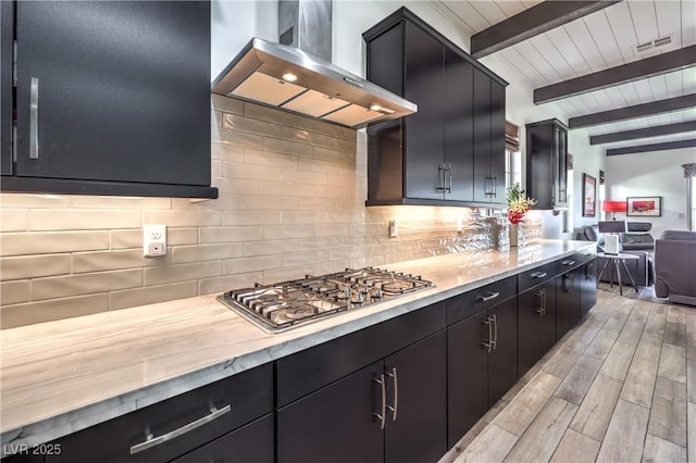 kitchen featuring dark cabinets, backsplash, wall chimney range hood, visible vents, and beamed ceiling