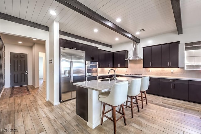 kitchen featuring light wood finished floors, stainless steel appliances, wall chimney range hood, a sink, and beam ceiling