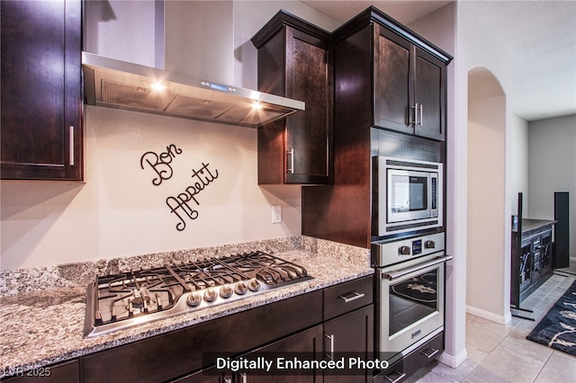 10470 Irish Cliffs Ct, Las Vegas, NV 89149 - kitchen featuring light stone countertops, arched walkways, appliances with stainless steel finishes, dark brown cabinets, and wall chimney exhaust hood