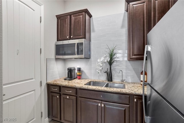kitchen featuring light stone counters, dark brown cabinets, a sink, backsplash, and appliances with stainless steel finishes