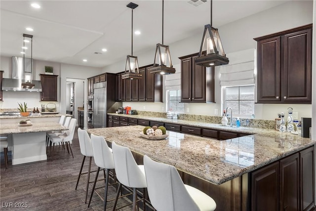kitchen featuring a kitchen breakfast bar, a sink, dark brown cabinets, wall chimney range hood, and light stone countertops
