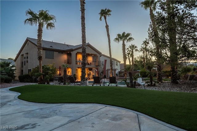 back of property at dusk with stucco siding, a tiled roof, a yard, and a balcony