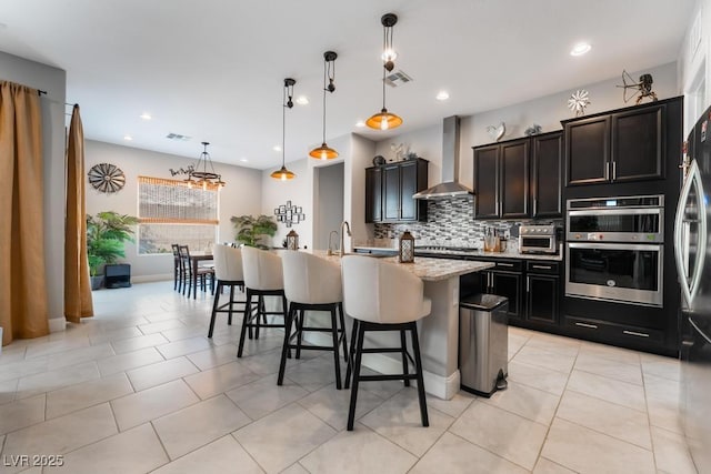 kitchen featuring a kitchen breakfast bar, visible vents, wall chimney range hood, an island with sink, and appliances with stainless steel finishes