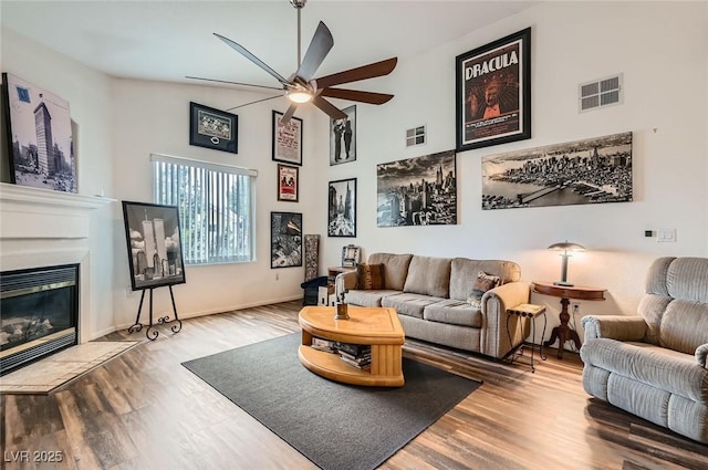 living room with wood finished floors, a glass covered fireplace, ceiling fan, a high ceiling, and visible vents