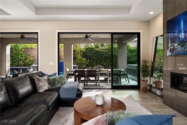 living room with a tiled fireplace, a ceiling fan, a tray ceiling, and recessed lighting