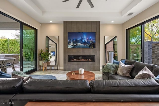 living room with a raised ceiling, wood finished floors, visible vents, a ceiling fan, and a tiled fireplace