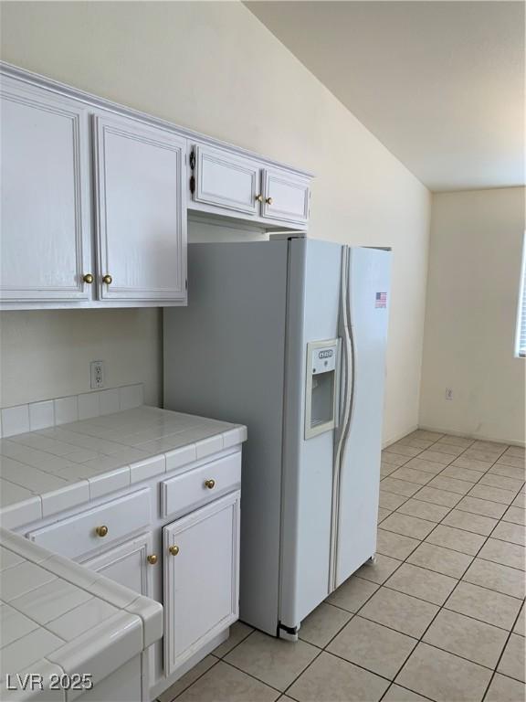 kitchen with white refrigerator with ice dispenser, tile countertops, light tile patterned floors, white cabinets, and lofted ceiling