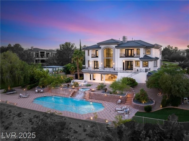 back of house at dusk with a tiled roof, a chimney, stairway, a balcony, and an in ground hot tub