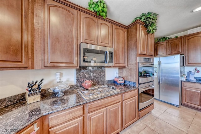 2733 Auchmull St, Henderson, NV 89044 - kitchen featuring stainless steel appliances, dark stone counters, light tile patterned floors, brown cabinetry, and a textured ceiling