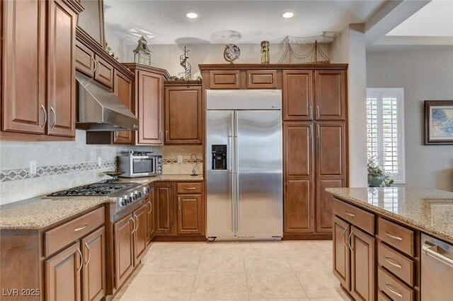 kitchen featuring light stone counters, decorative backsplash, light tile patterned floors, stainless steel appliances, and under cabinet range hood