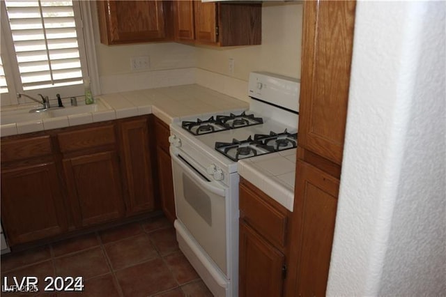 kitchen featuring brown cabinets, white range with gas stovetop, tile countertops, a sink, and dark tile patterned floors