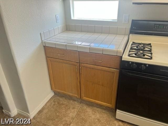 kitchen featuring baseboards, range with gas stovetop, brown cabinets, tile counters, and light tile patterned floors