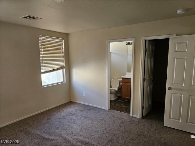 unfurnished bedroom with ensuite bathroom, baseboards, visible vents, dark carpet, and a walk in closet