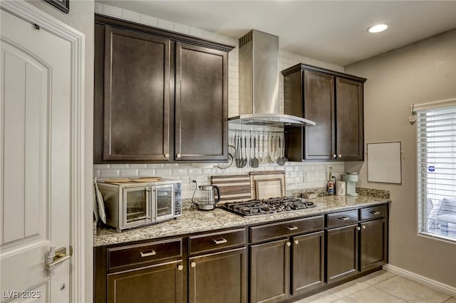 kitchen featuring stainless steel gas cooktop, dark brown cabinets, wall chimney range hood, light tile patterned floors, and backsplash