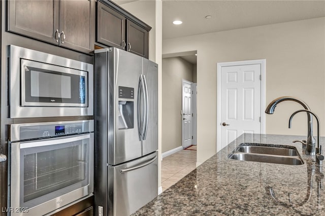 kitchen featuring stainless steel appliances, a sink, dark stone countertops, dark brown cabinets, and light tile patterned floors