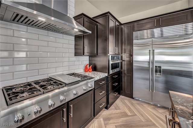 kitchen featuring tasteful backsplash, dark brown cabinets, stainless steel appliances, wall chimney exhaust hood, and light stone countertops