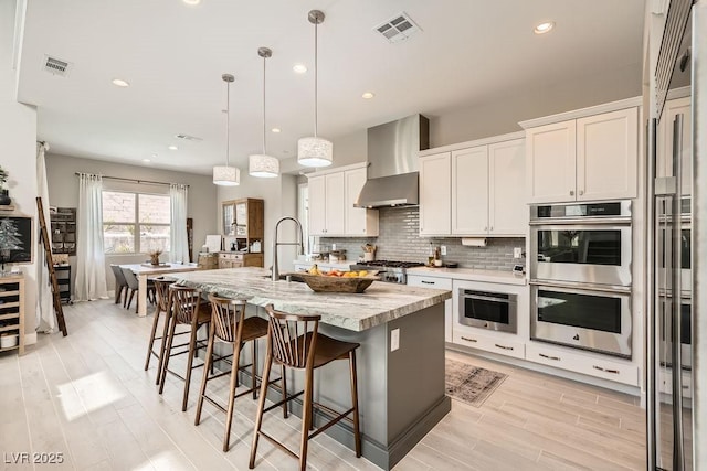 kitchen with wall chimney range hood, decorative backsplash, visible vents, a center island with sink, and double oven