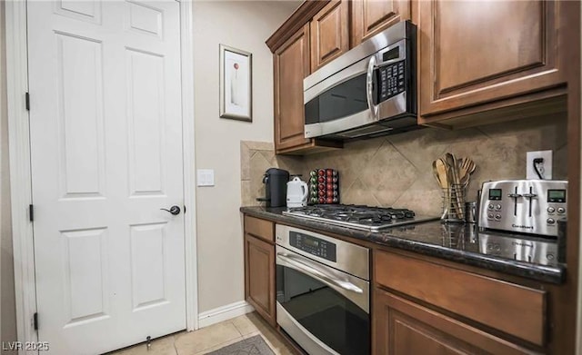 kitchen featuring dark stone countertops, decorative backsplash, brown cabinets, light tile patterned flooring, and stainless steel appliances
