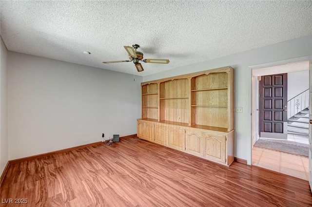 empty room featuring ceiling fan, stairs, baseboards, light wood-type flooring, and a textured ceiling