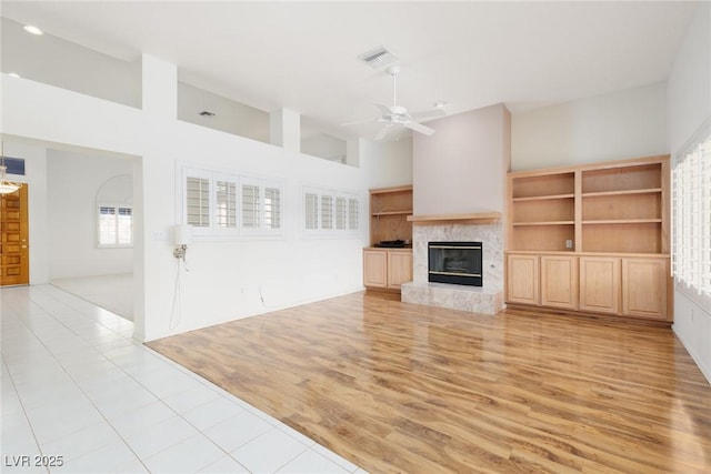unfurnished living room with visible vents, a glass covered fireplace, a high ceiling, a ceiling fan, and light wood-style flooring
