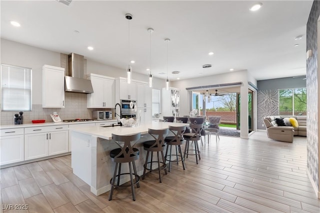 kitchen with decorative backsplash, gas stovetop, a center island with sink, light countertops, and wall chimney exhaust hood