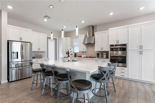 kitchen with wall chimney exhaust hood, stainless steel appliances, tasteful backsplash, visible vents, and light wood-style floors
