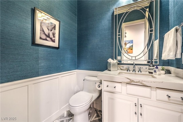 half bathroom with a decorative wall, wallpapered walls, wainscoting, vanity, and toilet