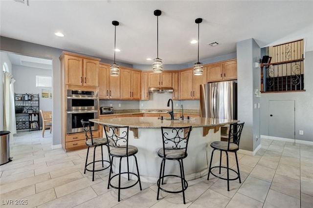 kitchen with under cabinet range hood, stainless steel appliances, light stone counters, light tile patterned floors, and a center island with sink