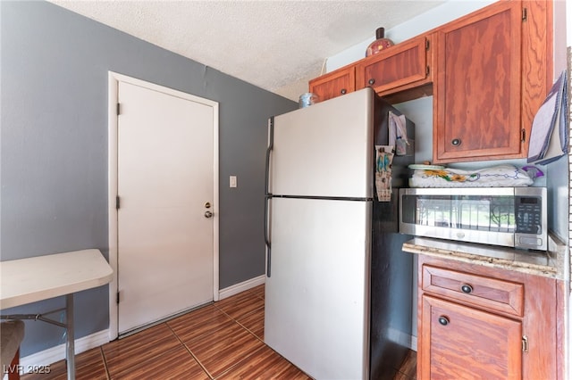 kitchen with freestanding refrigerator, stainless steel microwave, baseboards, a textured ceiling, and brown cabinets