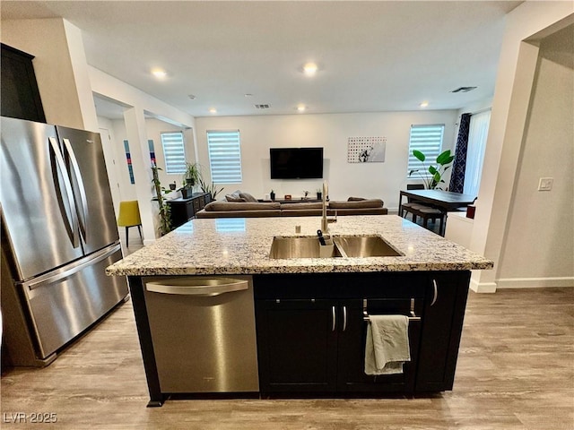 kitchen featuring stainless steel appliances, a sink, dark cabinets, light wood-style floors, and light stone countertops
