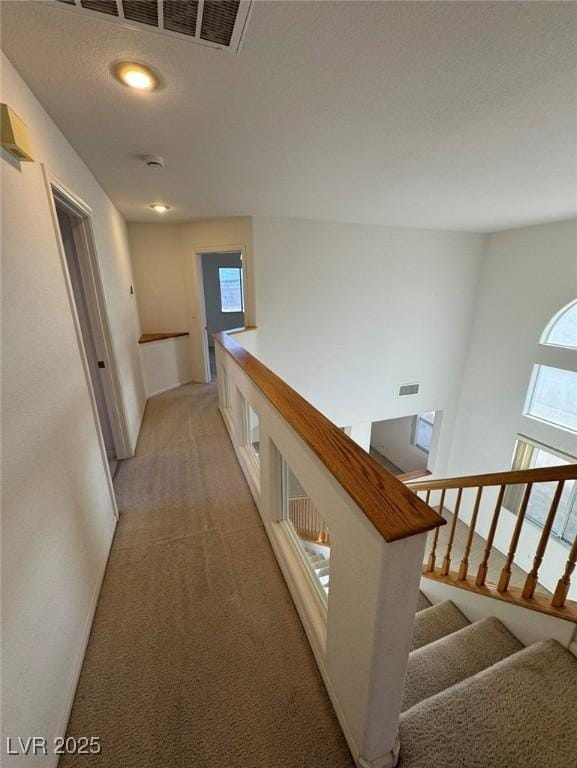 hallway with an upstairs landing, visible vents, light carpet, and plenty of natural light