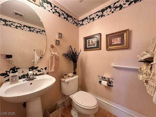 half bathroom with toilet, wallpapered walls, visible vents, and wood finished floors