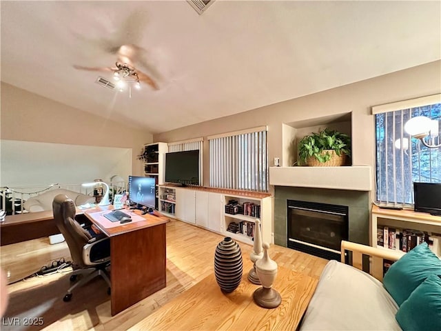 home office with visible vents, a glass covered fireplace, wood finished floors, a ceiling fan, and vaulted ceiling