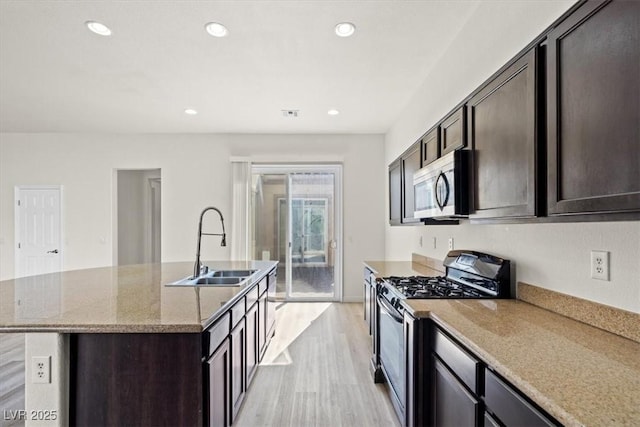 kitchen with a sink, stainless steel appliances, a center island with sink, dark brown cabinetry, and light wood-style flooring
