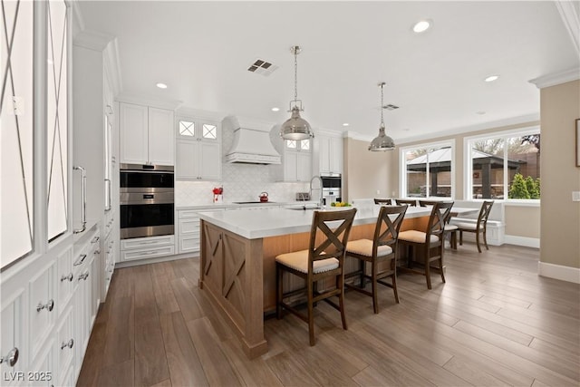 kitchen with black electric stovetop, custom range hood, double oven, visible vents, and a center island with sink