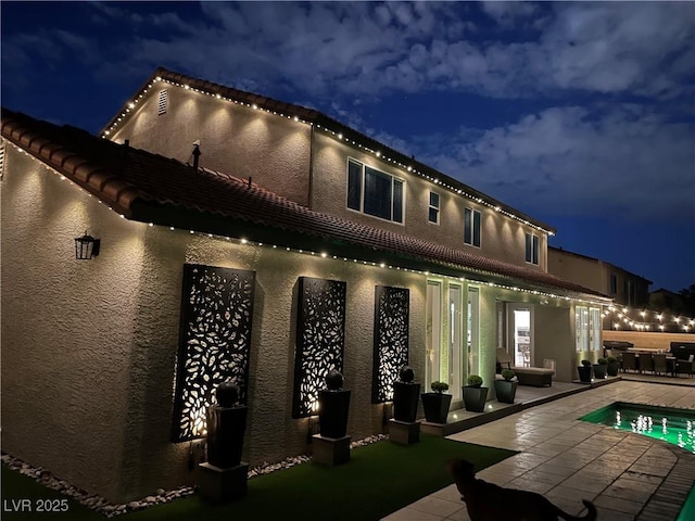 back of house at night featuring stucco siding, a patio area, and a fenced in pool