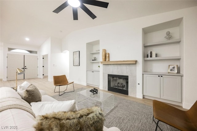 living room featuring light wood-style flooring, ceiling fan, a tile fireplace, vaulted ceiling, and built in features