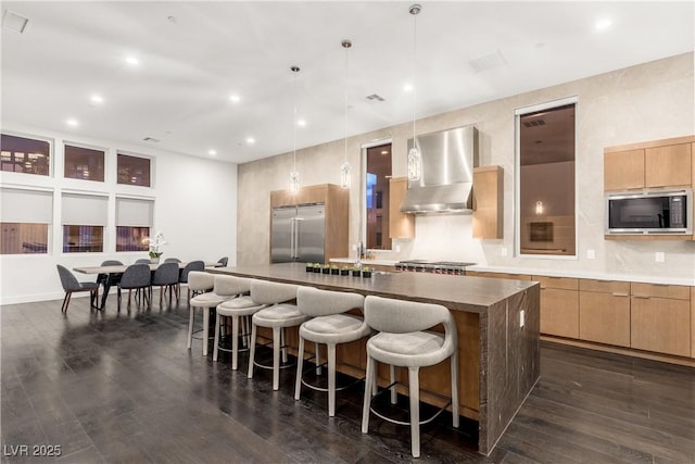 kitchen with a spacious island, a breakfast bar area, wall chimney range hood, built in appliances, and dark wood-style floors