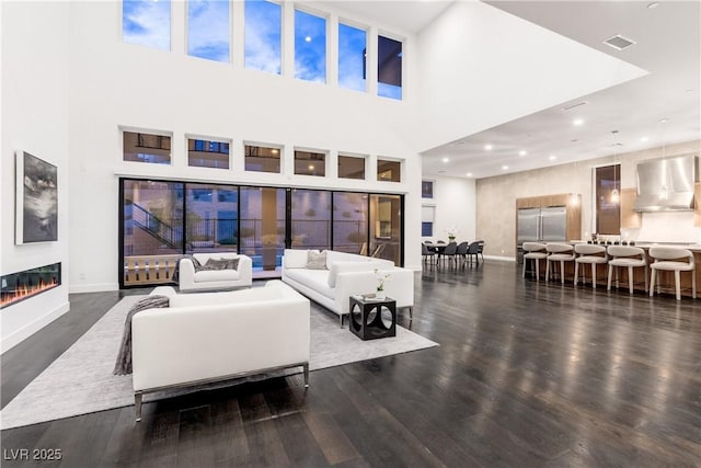 living room with dark wood finished floors, a glass covered fireplace, baseboards, visible vents, and a high ceiling