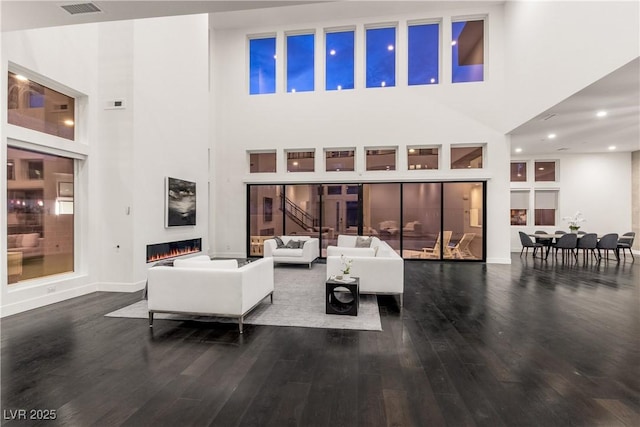 living area with a high ceiling, a glass covered fireplace, visible vents, baseboards, and wood finished floors