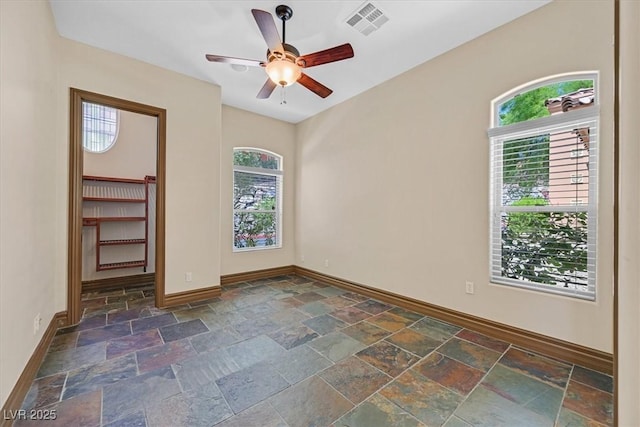 empty room featuring a ceiling fan, visible vents, stone tile flooring, and baseboards
