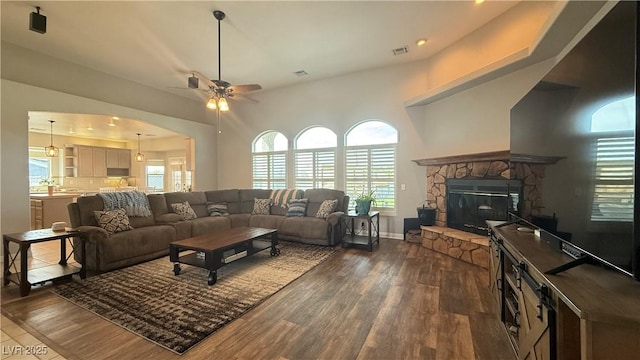 living room featuring a stone fireplace, ceiling fan, visible vents, and dark wood-style floors