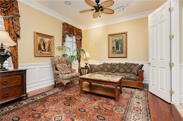 living area featuring ceiling fan, crown molding, a decorative wall, wainscoting, and wood finished floors
