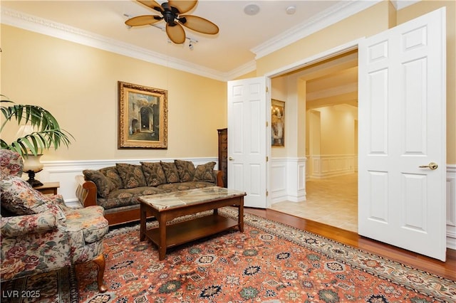 living area with a ceiling fan, a decorative wall, ornamental molding, wainscoting, and wood finished floors