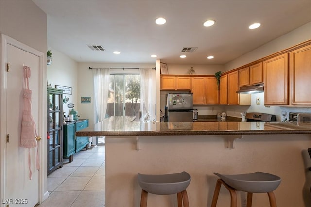 kitchen featuring visible vents, brown cabinets, light tile patterned floors, freestanding refrigerator, and under cabinet range hood