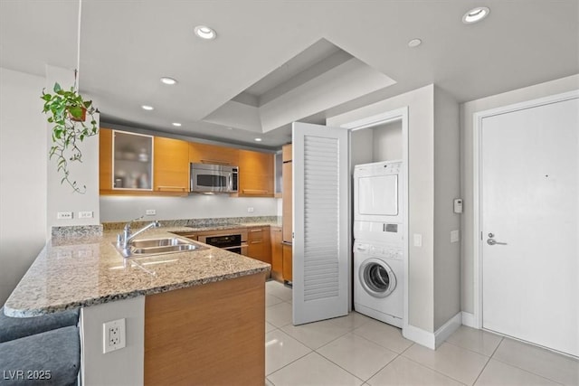 kitchen featuring a sink, appliances with stainless steel finishes, stacked washing maching and dryer, a peninsula, and brown cabinetry