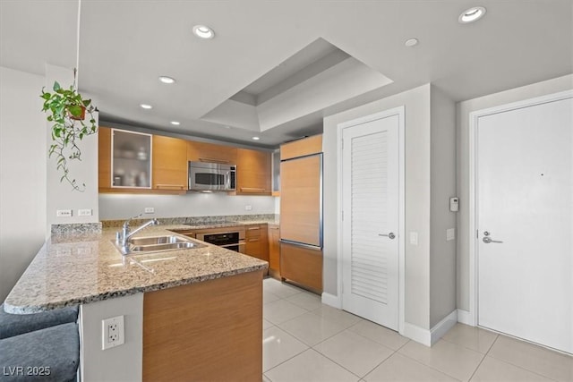 kitchen with light stone counters, a sink, appliances with stainless steel finishes, a peninsula, and brown cabinetry