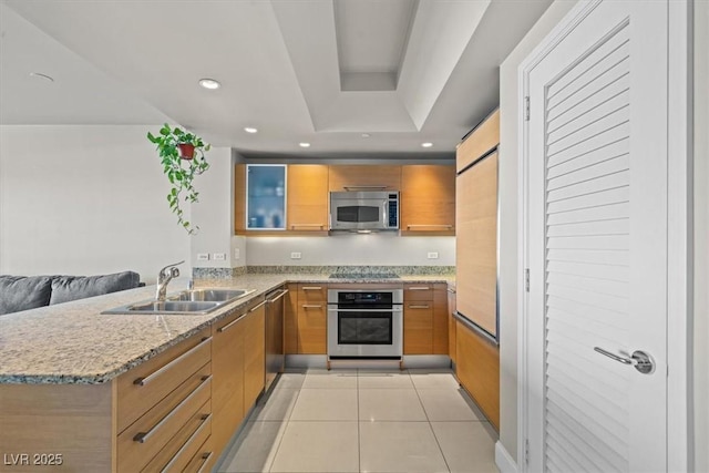kitchen featuring light stone counters, a sink, appliances with stainless steel finishes, a peninsula, and brown cabinetry