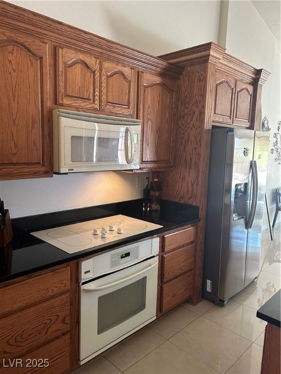 kitchen with brown cabinets, light tile patterned floors, dark countertops, and white appliances